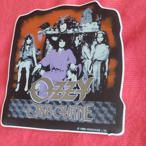 Pode incluir: Um autocolante com um gráfico de Ozzy Osbourne e outras figuras. A imagem tem uma borda preta e um fundo laranja e preto. O texto "Ozzy Osbourne" está em uma fonte estilizada. O autocolante está em uma superfície vermelha.