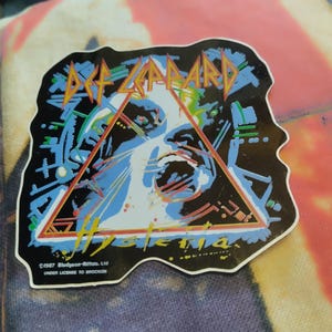 以下が含まれることがあります： Def Leppardのアルバム「Hysteria」のカバーアートが描かれたダイカットステッカー。中央の三角形、青と白のスタイライズされた顔、Def LeppardとHysteriaの文字がデザインされています。下部には1987 Bludgeon-Riffola, Ltd.という文字が記載されています。