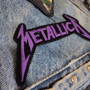 Könnte beinhalten: Nahaufnahme einer Jeansjacke mit mehreren Aufnähern. Der auffälligste Aufnäher zeigt den Bandnamen "METALLICA" in lila gestickten Buchstaben mit schwarzem Rand. Weitere Aufnäher sind sichtbar, darunter ein dreieckiger mit gelbem und grünem Design.