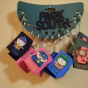 May include: A set of six South Park character keychains. Each keychain features a different character from the show, including Stan, Cartman, Kyle, Kenny, and others. The keychains are made of a soft, colorful material and are attached to silver key rings.