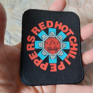 Può includere: Una toppa rettangolare in tessuto nero con una grafica centrale di un volto stilizzato rosso e grigio simile a un sole, circondato da una forma a stella azzurra. Il testo RED HOT CHILI PEPPERS è stampato in lettere rosse in cerchio.