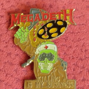 Può includere: Uno spillo in metallo dorato con un'immagine di un cartone animato di uno scheletro che indossa una maschera verde e un berretto da infermiera bianco. Lo scheletro tiene un set di batteria con la parola "MEGADETH" sopra. Il testo "KILLING IS MY BUSINESS" è su uno stendardo sotto lo scheletro.