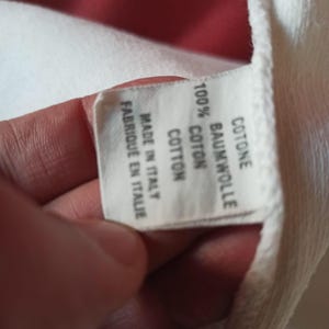 May include: A white fabric label with text in multiple languages, including "100% COTTON" and "MADE IN ITALY". The label is held by a hand, with a red background visible.