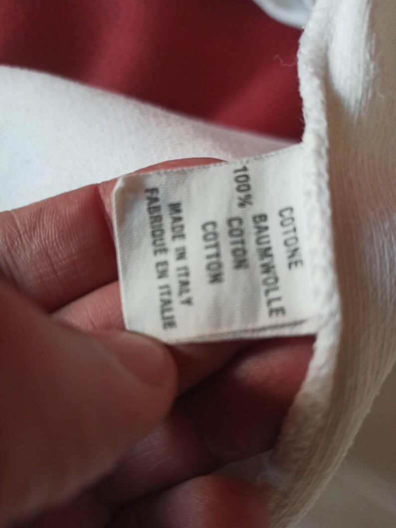 May include: A white fabric label with text in multiple languages, including "100% COTTON" and "MADE IN ITALY". The label is held by a hand, with a red background visible.