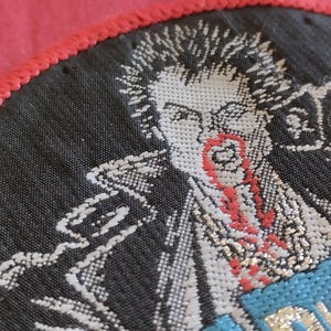 May include: Embroidered patch featuring a punk rock figure with spiky hair and a red, stylized blood-like design on the face and neck. The patch is black with white and red details, and a blue section with text. The patch is bordered by a red stitched edge.