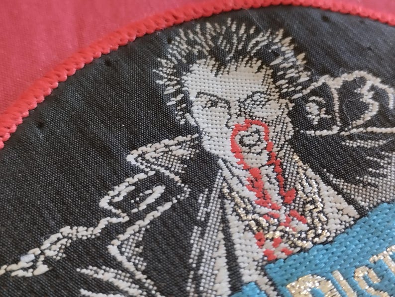 May include: Embroidered patch featuring a punk rock figure with spiky hair and a red, stylized blood-like design on the face and neck. The patch is black with white and red details, and a blue section with text. The patch is bordered by a red stitched edge.