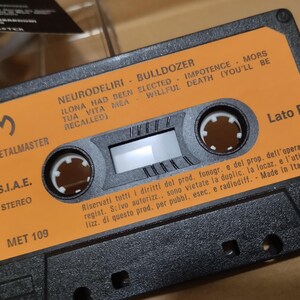 以下が含まれることがあります： オレンジ色のラベルが付いた黒いカセットテープ。ラベルには「Neurodeliri - Bulldozer」と「Ilona had been elected - Impotence - Mors tua vita mea - Willful death (you'll be recalled)」と書かれています。テープには「Metalmaster」と「S.I.A.E. Stereo」のラベルが付いています。テープには「Met 109」と「Lato B」のラベルが付いています。