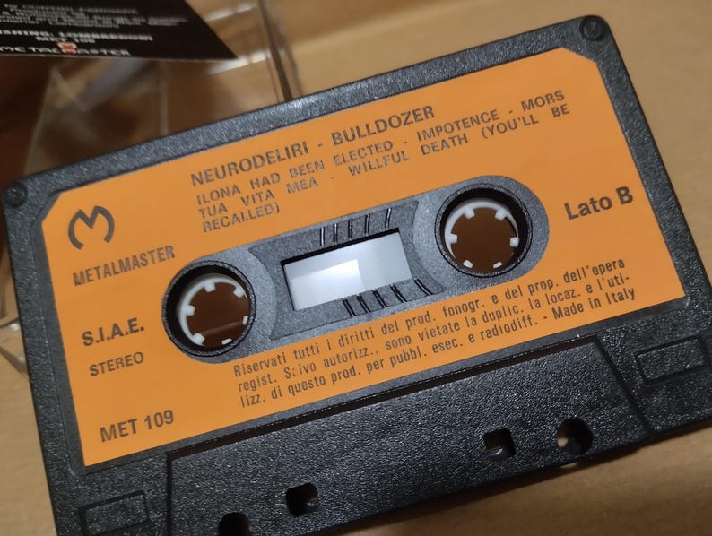 以下が含まれることがあります： オレンジ色のラベルが付いた黒いカセットテープ。ラベルには「Neurodeliri - Bulldozer」と「Ilona had been elected - Impotence - Mors tua vita mea - Willful death (you'll be recalled)」と書かれています。テープには「Metalmaster」と「S.I.A.E. Stereo」のラベルが付いています。テープには「Met 109」と「Lato B」のラベルが付いています。