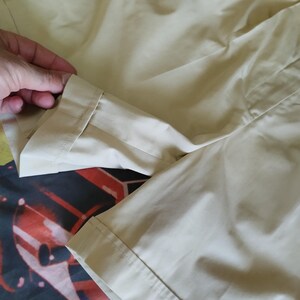 Puede incluir: Primer plano de tela color beige, posiblemente una prenda. La tela es lisa y parece ligera. La imagen muestra un dobladillo doblado y detalles de costura. El fondo es un patr&oacute;n colorido.