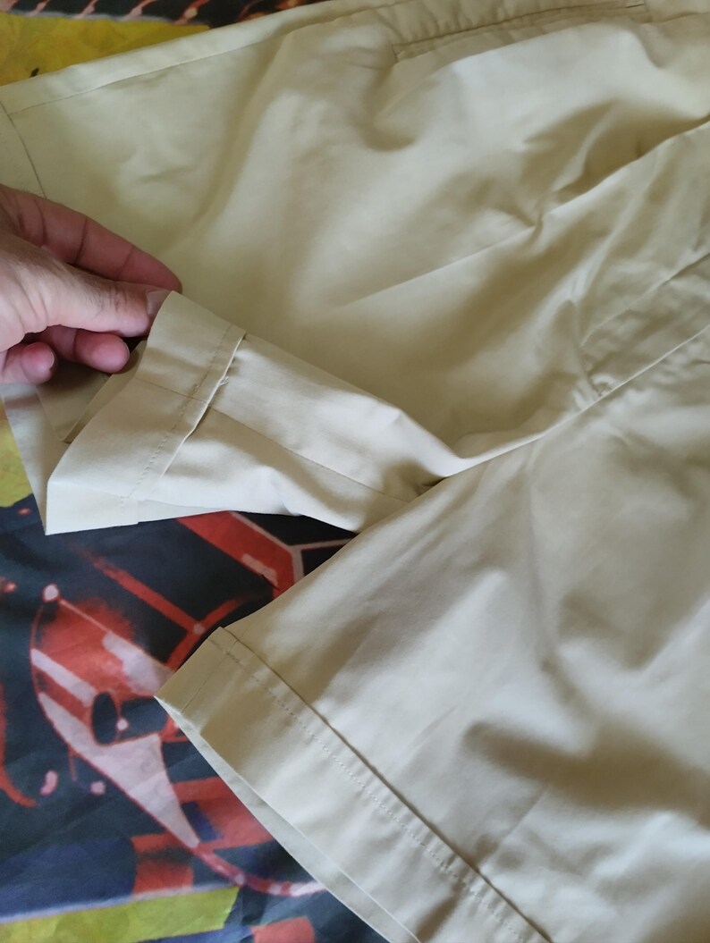 Puede incluir: Primer plano de tela color beige, posiblemente una prenda. La tela es lisa y parece ligera. La imagen muestra un dobladillo doblado y detalles de costura. El fondo es un patr&oacute;n colorido.