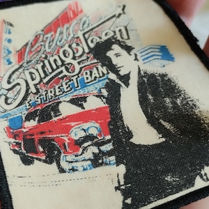 Puede incluir: Un parche de tela cuadrado con un borde cosido en negro que presenta un estampado gráfico de Bruce Springsteen y la E Street Band. El diseño incluye un coche vintage rojo, un edificio estilizado y el texto Bruce Springsteen E Street Band.