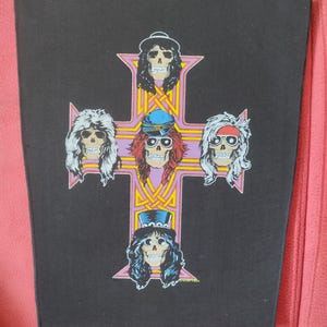 Puede incluir: Un parche negro con una cruz rosa y cinco caras de calavera de músicos. La cruz tiene un diseño amarillo y dorado. Los músicos llevan sombreros y tienen el pelo largo.