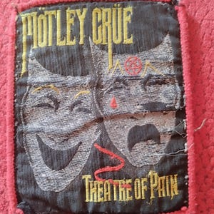 Könnte beinhalten: Ein schwarzer und grauer Aufnäher mit den Worten "MOTLEY CRUE" in gelben Buchstaben über zwei Theatermasken. Der Satz "THEATRE OF PAIN" steht unter den Masken. Der Aufnäher hat einen roten Rand.