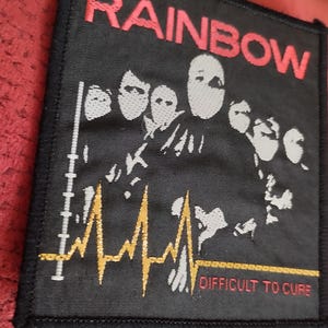 Könnte beinhalten: Schwarzer quadratischer Aufnäher mit dem Wort "RAINBOW" in roten Blockbuchstaben. Der Aufnäher zeigt eine weiße Grafik von mehreren maskierten Figuren und eine gelbe Herzschlaglinie. Der Text "DIFFICULT TO CURE" steht darunter.