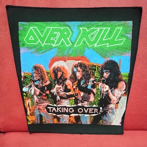 Op de afbeelding: Zwarte patch met de band Overkill, de tekst "Over Kill" in groen en "Taking Over" in wit. De afbeelding toont vier bandleden met lang haar, die instrumenten vasthouden, tegen een blauwe lucht en palmbomen.