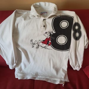 May include: White long-sleeved shirt with a Goofy graphic and the number 8 on the sleeve. The shirt has a collared neckline and button closure. The Goofy graphic is in red and black.