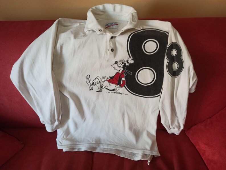 May include: White long-sleeved shirt with a Goofy graphic and the number 8 on the sleeve. The shirt has a collared neckline and button closure. The Goofy graphic is in red and black.