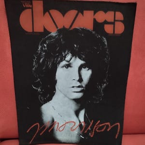 Peut inclure: Graphique en noir et blanc avec un portrait de Jim Morrison et le nom du groupe "The Doors" en orange en haut. Le nom "Morrison" est écrit en script rouge en dessous. L'image est sur fond noir.