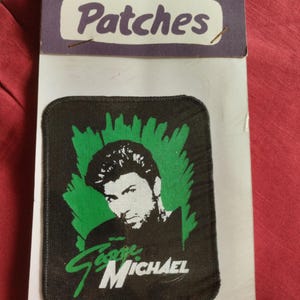 Op de afbeelding: Een zwarte patch met een portret van George Michael met een groene achtergrond. De patch heeft de naam "George Michael" in witte en groene letters. De patch is bevestigd aan een witte kaart met het woord "Patches" bovenaan.