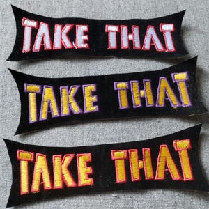 May include: Three black fabric patches, each shaped like a wide banner with pointed ends. Each patch features the embroidered text "TAKE THAT" in a bold, block font. The top patch has white text, the middle has yellow, and the bottom has red.