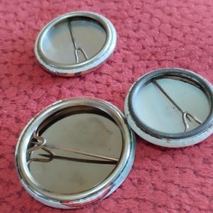 May include: Three blank button badges with silver metal pin backs. The badges are round and have a white background with a silver metal rim.