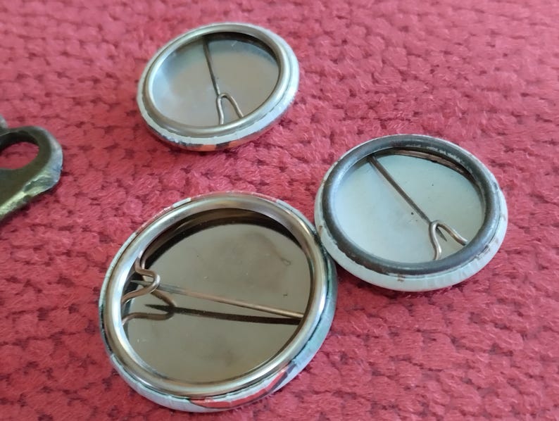 May include: Three blank button badges with silver metal pin backs. The badges are round and have a white background with a silver metal rim.