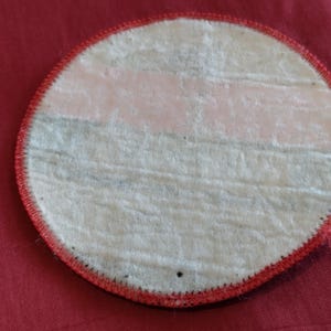 May include: A round patch with a white center and a red stitched border. The back of the patch has a light pink and gray striped design. The patch is on a red fabric surface.