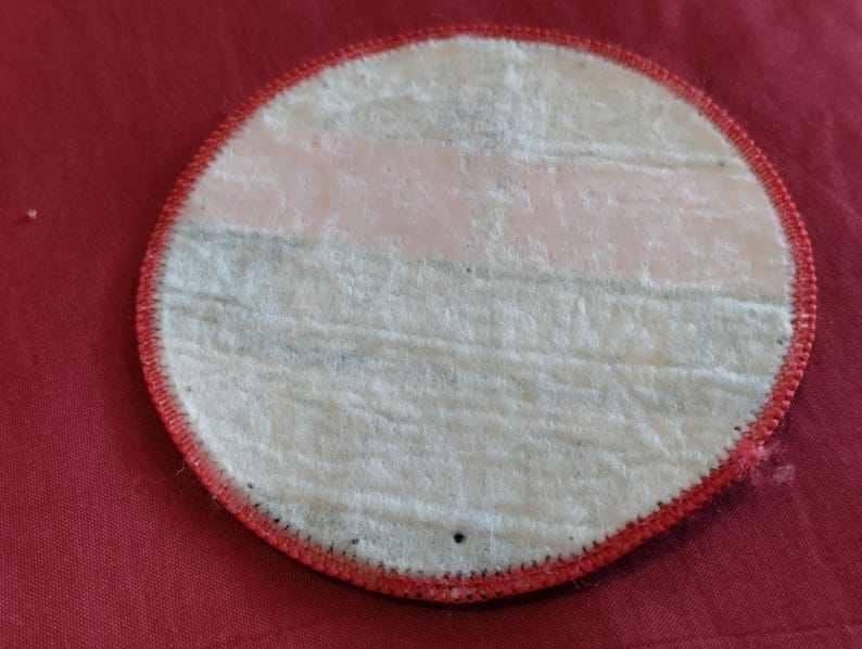 May include: A round patch with a white center and a red stitched border. The back of the patch has a light pink and gray striped design. The patch is on a red fabric surface.