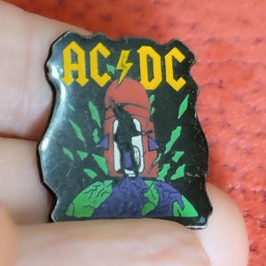 May include: A black AC/DC pin with a yellow logo and lightning bolt. The pin features a red and black graphic of a figure on a tower over a purple and green globe. The pin is held in a hand.