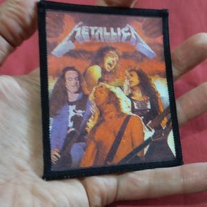 Può includere: Una toppa rettangolare con bordo nero, raffigurante la band Metallica. La toppa mostra un'immagine colorata dei membri della band. Il nome della band, "Metallica", è stampato in lettere bianche in alto.