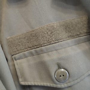 May include: A gray utility jacket with a large pocket on the front. The pocket has a flap with a velcro closure and a button.