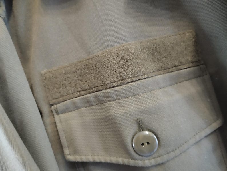 May include: A gray utility jacket with a large pocket on the front. The pocket has a flap with a velcro closure and a button.