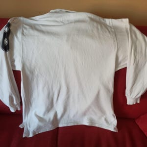 May include: A white long-sleeved shirt with a crew neck. The left sleeve has a black design. The shirt is made of a soft material, possibly cotton. The shirt is laid out on a red surface.