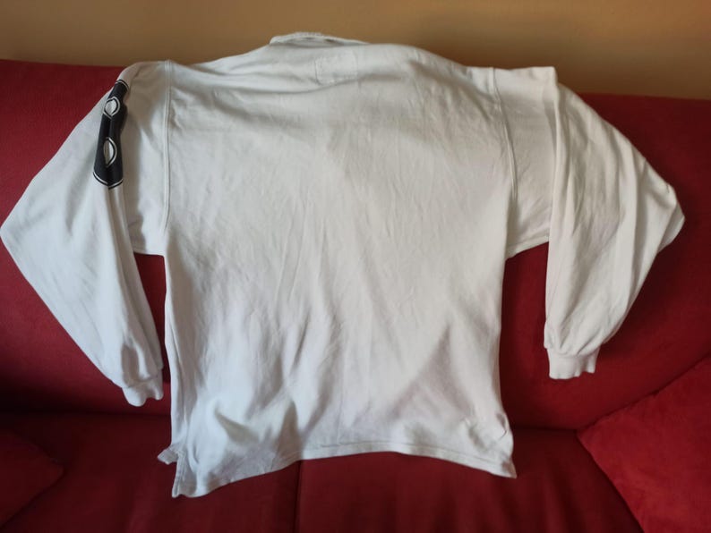 May include: A white long-sleeved shirt with a crew neck. The left sleeve has a black design. The shirt is made of a soft material, possibly cotton. The shirt is laid out on a red surface.