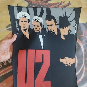 Peut inclure: Un patch en tissu noir et rouge avec le logo d'un groupe. Le logo est le mot "U2" en lettres rouges. Quatre hommes sont représentés en noir et blanc derrière le logo.