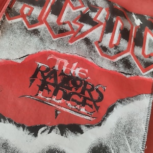 Könnte beinhalten: Ein rot-weißes Grafikdesign mit dem Bandnamen "AC/DC" in roten Blockbuchstaben. Darunter steht der Text "The Razor's Edge" in einer gotischen Schriftart auf rotem Hintergrund.