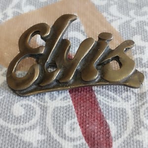 May include: A gold-colored metal pin with the word "Elvis" written in a stylized script font.