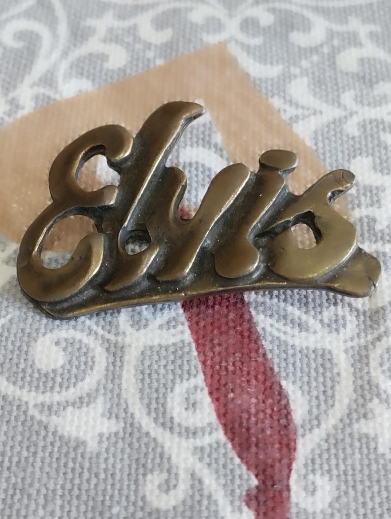 May include: A gold-colored metal pin with the word "Elvis" written in a stylized script font.