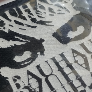 May include: A close-up of a black and white textile featuring the word "BAUHAUS" in a stylized font. The design has a bold, graphic style, with the letters partially obscured by a dark, abstract shape.