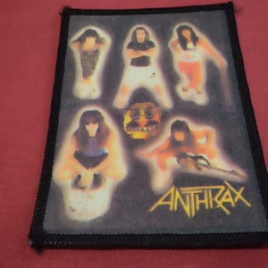 Op de afbeelding: Een rechthoekige stoffen patch met een zwarte gestikte rand. Het ontwerp toont vijf figuren en een centraal gezicht op een donkergrijze achtergrond. Het woord ANTHRAX staat in gele hoofdletters in de rechterbenedenhoek gedrukt.