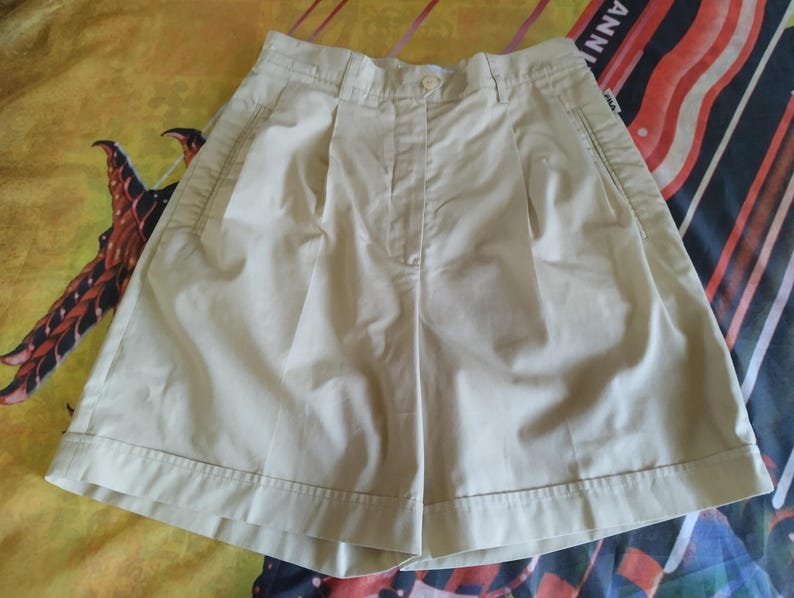 Puede incluir: Pantalones cortos color crema con cierre de bot&oacute;n y pliegues. Los pantalones cortos tienen bolsillos laterales y un ajuste a medida. La tela parece ser un algod&oacute;n ligero o mezcla, adecuado para climas c&aacute;lidos.