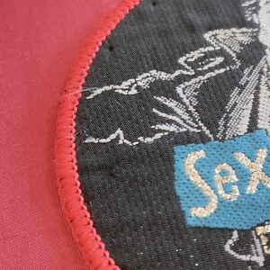 May include: A round, black patch with a red stitched border. The patch features a design with white and red accents, and the words "Sex Pistols" in a light blue box with gold lettering.