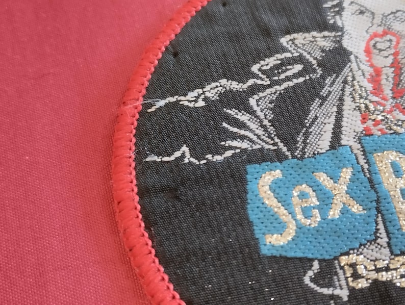 May include: A round, black patch with a red stitched border. The patch features a design with white and red accents, and the words "Sex Pistols" in a light blue box with gold lettering.