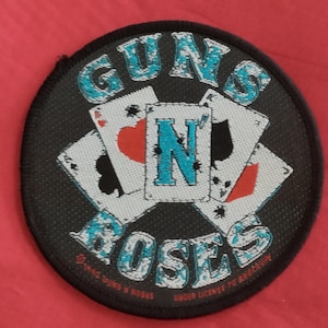 May include: A circular black patch with the band name "GUNS N' ROSES" in turquoise lettering. The design includes playing cards and the text "UNDER LICENSE TO BROOKLYN" and "DANGEROUS N' ROSES".