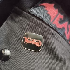 May include: A black and red Norton pin on a black fabric background.