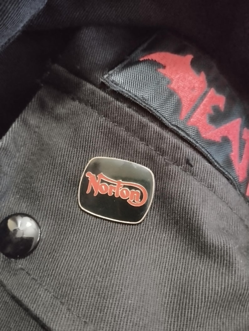 May include: A black and red Norton pin on a black fabric background.