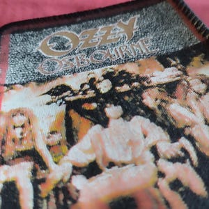 Könnte beinhalten: Ein rechteckiger Aufnäher mit dem goldenen Text "OZZY OSBOURNE". Der Aufnäher hat einen schwarzen Hintergrund mit einem verblassten Bild von Personen. Die Ränder sind rot und schwarz.