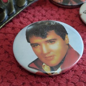 May include: A white button with a picture of Elvis Presley wearing a red jacket and a white shirt. The button has a white background and a black border.