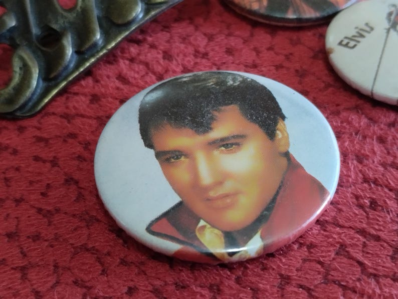 May include: A white button with a picture of Elvis Presley wearing a red jacket and a white shirt. The button has a white background and a black border.