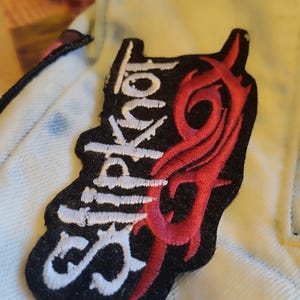 May include: A black patch featuring the word "Slipknot" in white, stylized lettering, and a red tribal design. The patch is sewn onto a light yellow fabric.